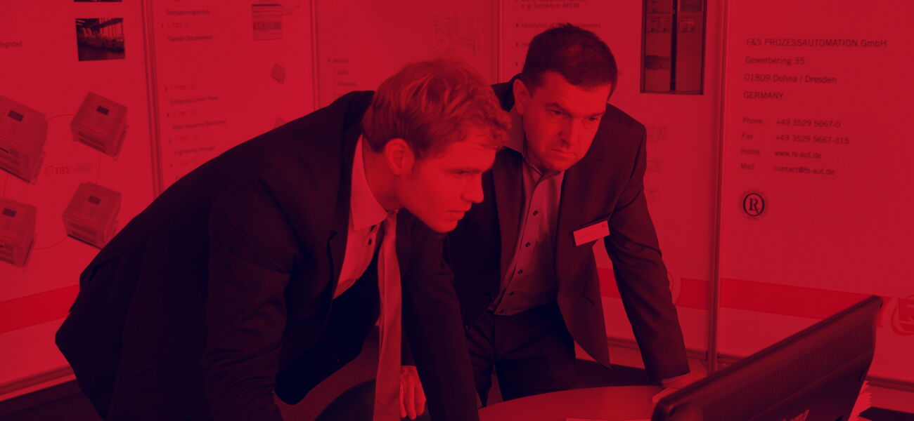 Rötliches Bild - Zwei Männer stehen an einem Tisch und schauen in einen Computer.